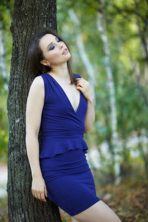Female in blue dress leaning against a tree trunk in autumn linden tree alley. Path under yellow trees with falling autumn leaves. Sunny autumn day.の写真素材