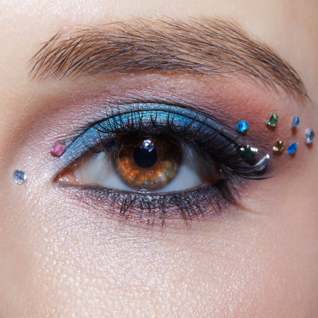 Closeup macro shot of human female brown color eye. Woman with natural evening vogue face beauty makeup. Girl with perfect skin, with blue eye shadow make up and rhinestones.の写真素材