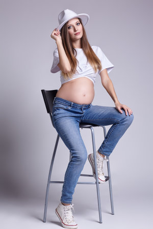 Young pretty pregnant woman in white t-shirt, and jeans sit on bar stool. Female with belly exposed. 5th month of pregnancy.の写真素材