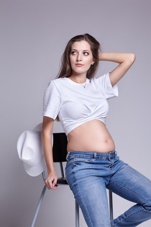 Young pretty pregnant woman in white t-shirt, and jeans sit on bar stool. Female with belly exposed. 5th month of pregnancy.の写真素材