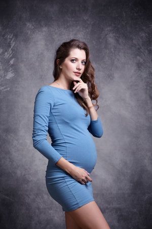 Portrait of young pretty pregnant woman on gray studio background. Female in blue dress with hands near pregnant belly.の写真素材
