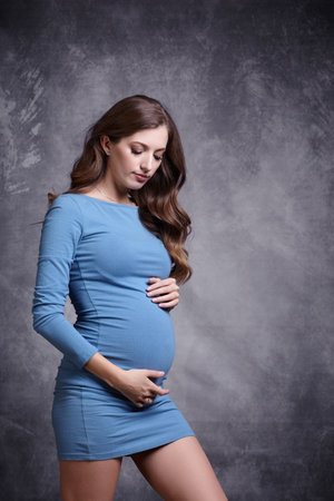 Portrait of young pretty pregnant woman in blue dress on gray studio background. Female looking at pregnant belly and supports the stomach with hands.の写真素材