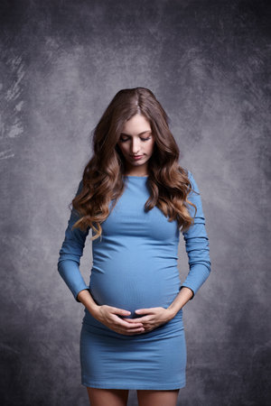 Portrait of young pretty pregnant woman in blue dress on gray studio background. Female looking at pregnant belly and supports the stomach with hands.の写真素材
