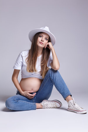 Young pretty pregnant woman in white t-shirt, hat and jeans sit on the floor on background. Female with belly exposed. 5th month of pregnancy.の写真素材