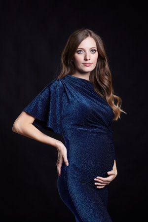 Portrait of young pretty pregnant woman on black studio background. Female in blue sequin dress. One hand on hip, another on the belly.の写真素材