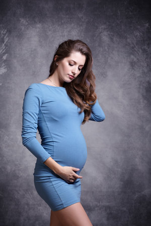 Portrait of young pretty pregnant woman in blue dress on gray studio background. Female looking at pregnant belly and supports the stomach with hands.の写真素材