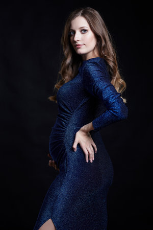 Portrait of young pretty pregnant woman on black studio background. Female in blue sequin dress, hand on hip.の写真素材