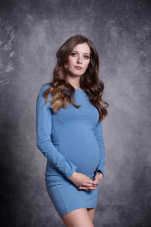 Portrait of young pretty pregnant woman on gray studio background. Female in blue dress with hands near pregnant belly.の写真素材