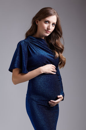 Portrait of young pretty pregnant woman on gray studio background. Female in blue sequin dress with hands near pregnant belly.の写真素材
