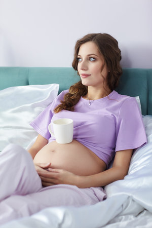 A pregnant woman lies in bed of her domestic bedroom. Female dressed in pajama enjoying her pregnancy state. A cup of tea is on the belly.の写真素材