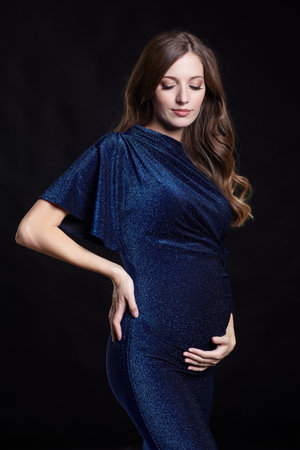 Portrait of young pretty pregnant woman on black studio background. Female in blue sequin dress. One hand on hip, another on the belly.の写真素材