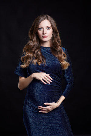 Portrait of young pretty pregnant woman on black studio background. Female in blue sequin dress with hands near pregnant belly.の写真素材
