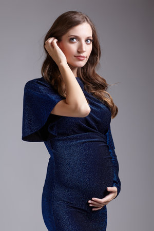 Portrait of young pretty pregnant woman on gray studio background. Female in blue sequin dress. One hand near pregnant belly, another near face.の写真素材
