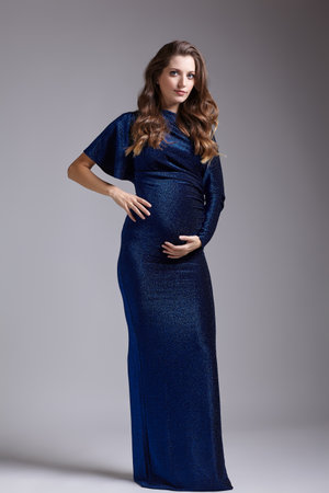 Portrait of young pretty pregnant woman on gray studio background. Female in blue sequin dress with hands near pregnant belly.の写真素材