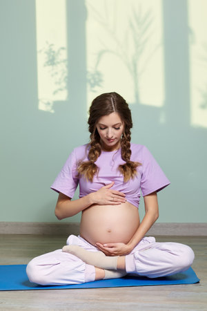 A pregnant woman with long brown hair sits cross-legged, meditating and practicing yoga during pregnancy. Woman looks at her pregnant belly. On the background are shadows of pot plants on the window.の写真素材