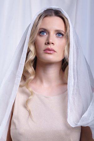 Portrait of a young blue eyed woman against a background of white fabric. The woman is wearing a cream beige dress with straps. Female head is covered with a white cloth.の写真素材