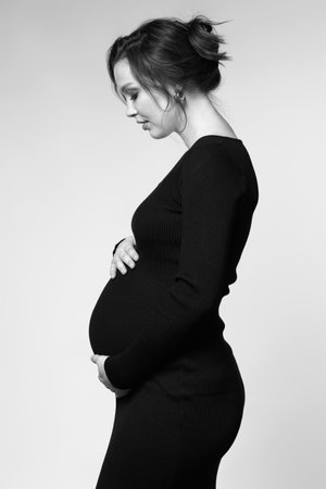 Black and white Portrait of young pretty pregnant woman on gray studio background. Female in black dress with hands near pregnant belly.の写真素材