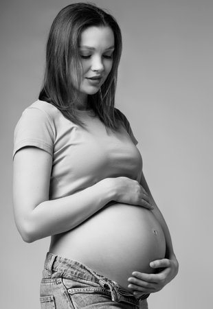 Black and white photo of Young pretty pregnant woman in t-shirt and jeans on gray background. Female...