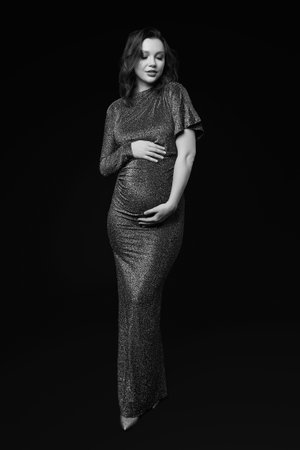Black and white portrait of pretty pregnant woman on black studio background. Female in gray sequin dress with hands near pregnant belly.の写真素材