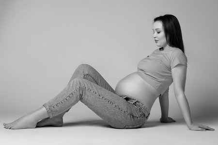 Black and white photo of young pretty pregnant woman in t-shirt and jeans sit on the floor on gray background. Female with belly exposed. 6th month of pregnancy.の写真素材