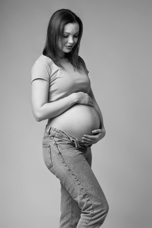 Black and white portrait of young pretty pregnant woman in t-shirt and jeans on gray background. Female looking at pregnant belly. Belly exposed. 6th month of pregnancy.の写真素材
