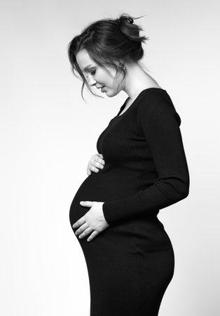 Black and white portrait of young pretty pregnant woman on gray studio background. Female in black dress with hands near pregnant belly.の写真素材