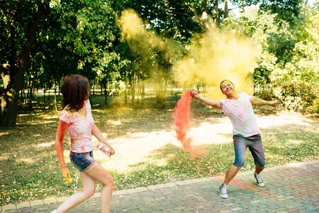 Young and beautiful, hipster couple funny playing in the park on holi color festival throw each other with colour paint powder. Dressed in white T-shirts and denim shorts.の写真素材