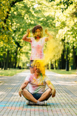 Young and beautiful, hipster couple funny playing in the park on holi color festival with colour paint powder. Dressed in white T-shirts and denim shorts. Girl throws paint on the guy's head.の写真素材