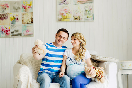 Couple dressed in blue and white colors with pregnant woman relaxing on white sofa in white room together. They are funny playing with bears.の写真素材