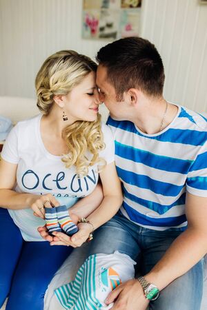 Young pregnant woman with husband on white sofa in white room lovely hold baby shoes. Couple dressed in blue and white colors.の写真素材