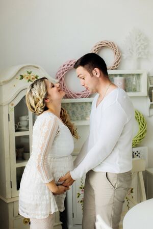Happy young pregnant wife with husband dressed in white clothes lovely touching belly at home. Sensual photo.の写真素材