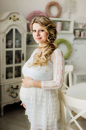 Beautiful pregnant woman in white dress caressing her belly in white room and looking at the camera. Sensual photo.の写真素材