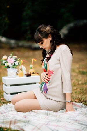 Beautiful pregnant woman is sitting and looking lovely on tummy on the checkered blanket with her hand on belly. Autumn picnic.の写真素材