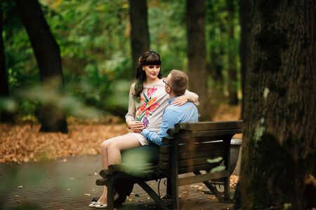 Beautiful pregnant stylish couple relaxing outside in the autumn park sitting on bench. They are lovely looking into each other's eyes.の写真素材