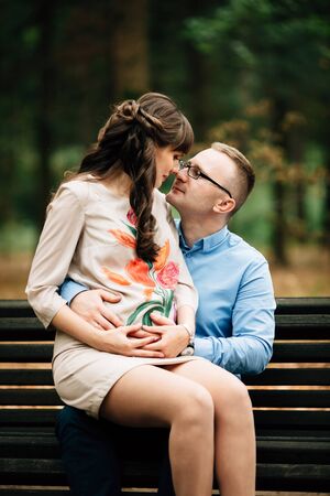Beautiful pregnant stylish couple relaxing outside in the autumn park sitting on bench. They are lovely looking into each other's eyes.の写真素材