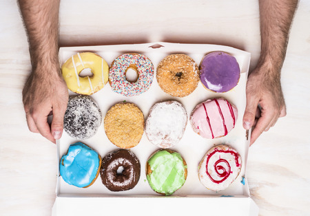 White paper box full of doughnuts in male hands on wooden background, top viewの写真素材