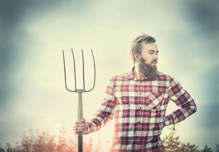 young bearded farmer in red checkered shirt with old pitchfork on sky  nature backgrund, tonedの写真素材