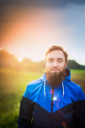 young man with a beard wearing a blue jacket and in profile against green field and skyの写真素材