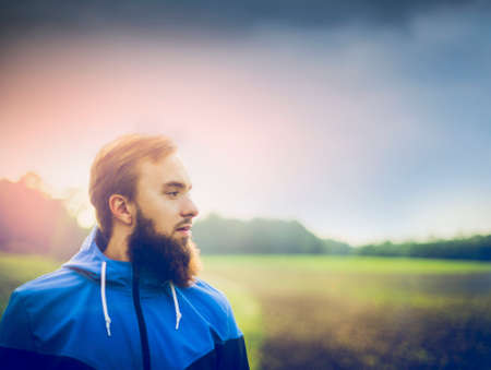 young man with a beard wearing a blue jacket and in profile against green field and skyの写真素材