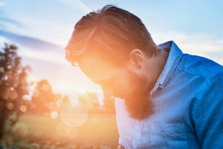 young man in a shirt with a beard stands on a sunny background trees in a fieldの写真素材
