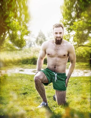 sporty young man with a naked torso and a beard, standing on one knee on the grass in the natural background of the sky and treesの写真素材