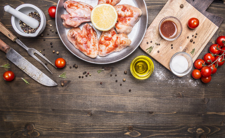 raw chicken wings with barbecue sauce with peppers, herbs, a knife and fork, butter, herbs, cherry tomatoes on wooden rustic background top view close up border, place for textの写真素材