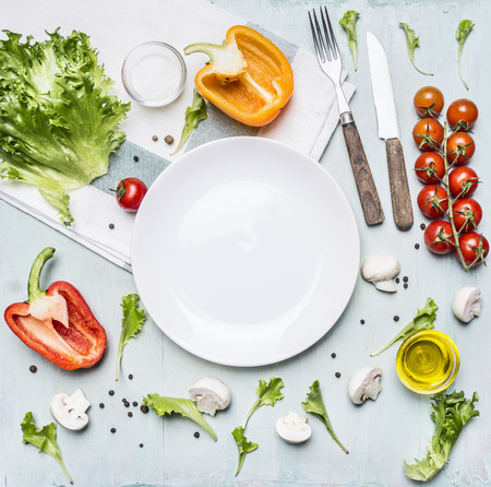 Ingredients for cooking salad cherry tomatoes, lettuce, peppers, spices and oil  laid out around a white plate on wooden rustic background top view close up