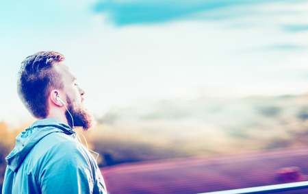 young man with a beard and a short haircut, standing in profile against a blurred urban sunny landscape with headphones in their earsの写真素材