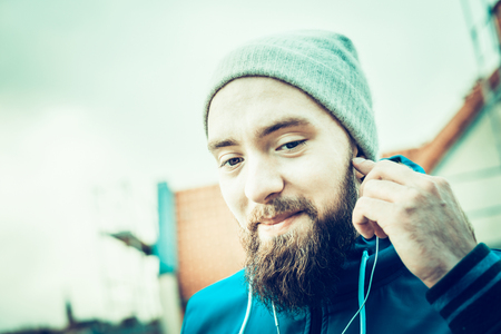 man with a beard, wearing a hat and smiling with headphones on the urban background blurred landscapeの写真素材