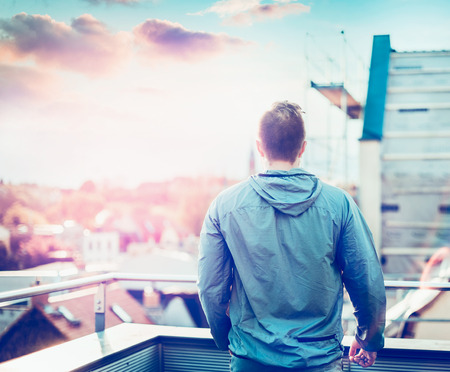 young man in gray jacket with short hair, standing on the balcony of the house and admires the sunset blurred cityscapeの写真素材