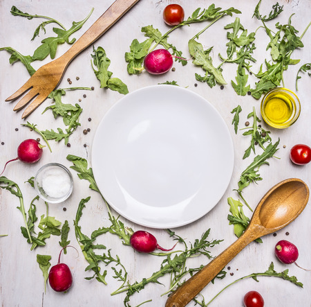 Healthy foods, cooking and vegetarian concept arugula salad, wooden spoon and fork for the salad, radishes, cherry tomatoes, seasonings and oils lined up around the white saucer, frame, space for textの写真素材