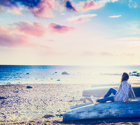 toned landscape, girl sitting on the beach on an inverted boat and looking at the sea, a beautiful evening skyの写真素材