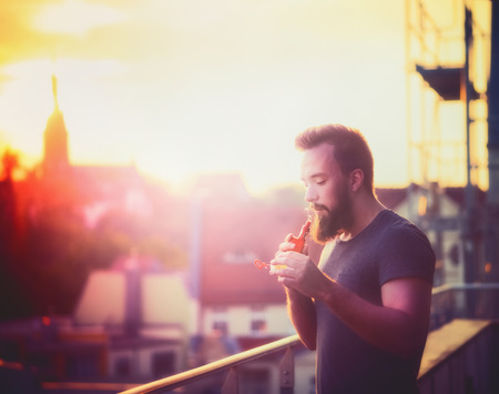 young man with a beard making soap bubbles with the help of Vaporizers against the evening urban landscape blurredの写真素材