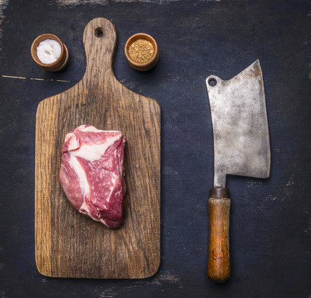 Fresh raw pork steak on a cutting board with vintage meat cleaver on a dark wooden background with pepper and saltの写真素材